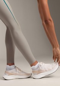 Scarpe da ginnastica bianche con tomaia in rete, dettagli beige e suola in gomma, abbinate a leggings attillati grigi chiari con strisce turchesi.