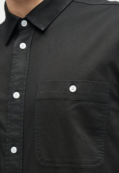 Μαύρο πουκάμισο με λευκά κουμπιά, που διαθέτει τσέπη στο στήθος ασφαλισμένη με ένα λευκό κουμπί, με γιακά ορατό σε πρόσωπο.