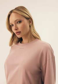 Jasnoróżowa koszula z długim rękawem, okrągłym dekoltem, z subtelnym logo na klatce piersiowej oraz gładką, miękką teksturą.