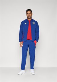Chándal deportivo azul con acentos rojos, chaqueta con cremallera, bolsillos con cremallera y rayas blancas. Se lleva con una camiseta roja y zapatillas blancas.