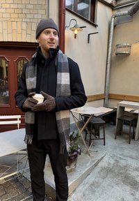 Sort frakke med et grå- og hvidternet tørklæde, brune handsker, holder en kaffekop, sat i en café med trækborge og en brostensgulv.