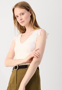 Top s véčkovým výstřihem v světle růžové barvě, s jemnými krajkovými detaily na ramenou, spárovaný s olivovými kalhotami s vysokým pasem a černým páskem.