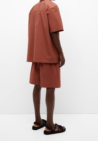 Kurzärmliges, strukturiertes, rostfarbenes Hemd mit passenden Shorts. Lockere Passform, minimale Details, kombiniert mit schwarzen Sandalen mit Riemchen.