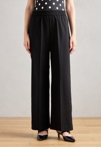 Black wide-leg pants with an elastic waistband, made from a smooth material. Worn with black heeled shoes, set against a neutral background.