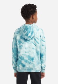 Chico con el pelo corto y rizado, vistiendo una sudadera con capucha de teñido anudado en azul y blanco, de espaldas, acompañado de pantalones negros, contra un fondo sencillo.