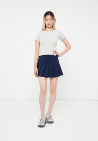 Világosszürke, rövid ujjú felső "Santa Cruz" grafikával, sötétkék, plisszírozott mini szoknyával és szürke, fekete részletekkel ellátott slip-on cipővel párosítva.