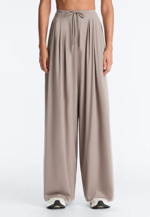 Vide ben i taupe med samlet talje og snøre, båret med hvide sneakers på en ensfarvet baggrund.