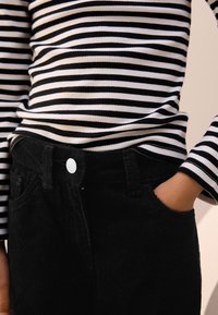 Schwarze Cordhosen mit einer frontalen Tasche und weißem Knopf, kombiniert mit einem schwarz-weiß gestreiften Langarmshirt mit geripptem Muster.