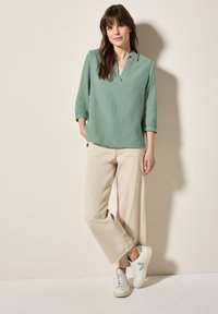 Maglietta verde menta a maniche lunghe con colletto e tessuto texture, abbinata a pantaloni beige larghi e sneakers bianche con dettagli blu.