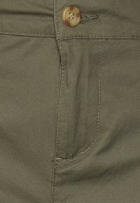 Detailní záběr na olivově zelené kalhoty s knoflíkovým pasem a precizním prošíváním kolem kapsy a zipu.
