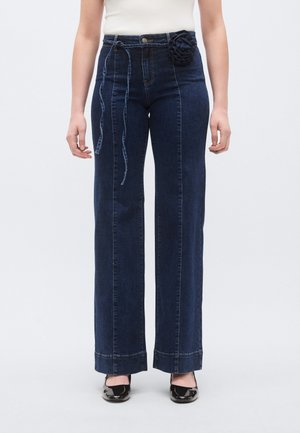 Vaqueros de mezclilla oscuros de tiro alto y pierna ancha, con una flor decorativa de tela en la cintura y costuras en contraste a lo largo de las costuras.