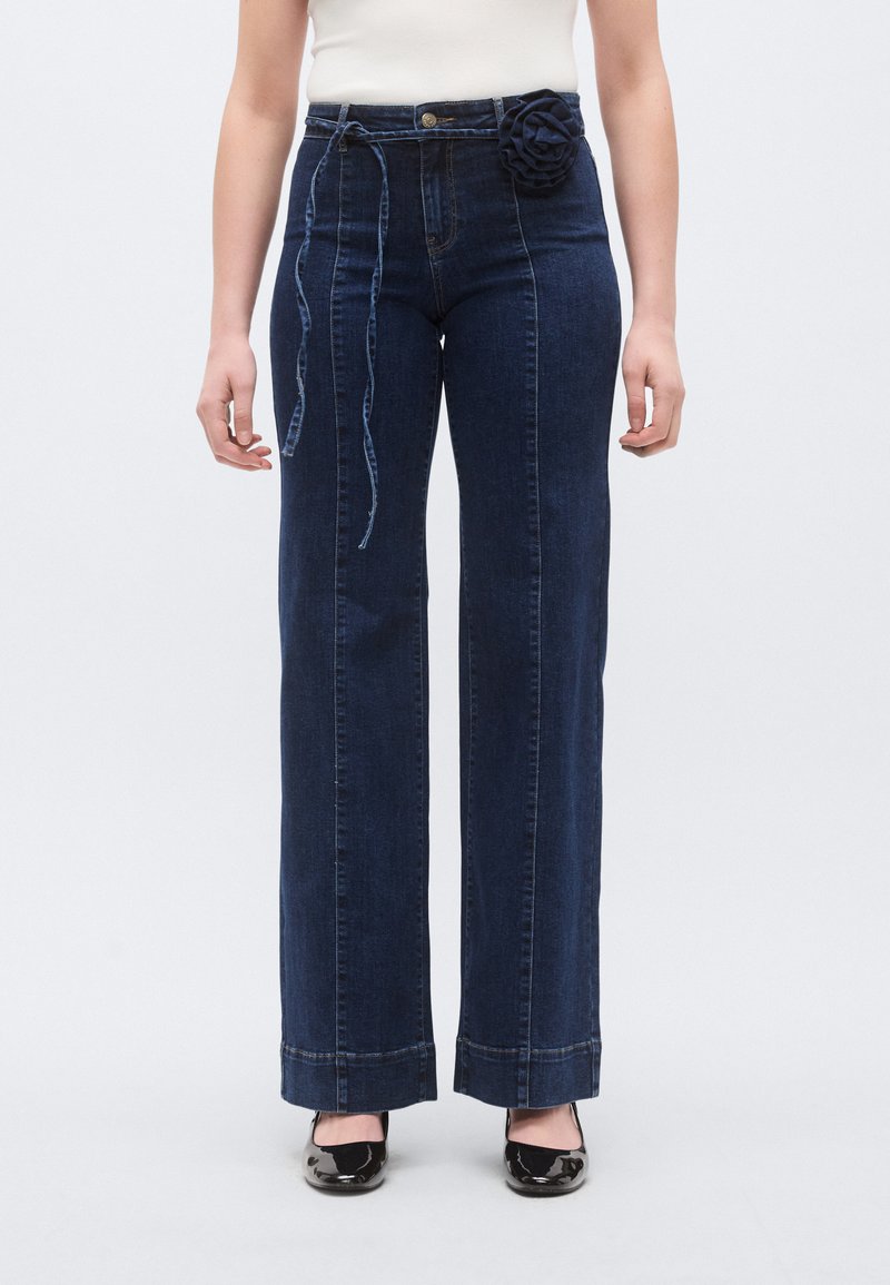 Τζιν παντελόνι με ψηλή μέση και φαρδιά πόδια σε σκούρο τζιν ύφασμα, διακοσμημένο με υφασμάτινο λουλούδι στη μέση και αντίθετη ραφή κατά μήκος των ραφών.