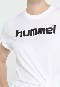 Bílý bavlněný tričko s výrazným černým logem "hummel" tištěným na hrudi. Klasický kulatý výstřih a krátké rukávy s ohrnutím.