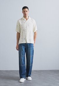Kortermet kremfarget skjorte med teksturerte utskjæringer, kombinert med løse blå denimjeans med lappesøm-design, og hvite joggesko.