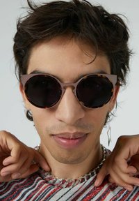 Lunettes de soleil rondes noires avec des accents en écaille de tortue, une monture épaisse et une finition mate, portées par une personne sur un fond clair.