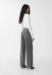 Bílý dlouhý rukáv crop top v kombinaci s vysokými šedými kalhotami širokého střihu s dvojitými zadními kapsami. Černé jehlové podpatky dotvářejí vzhled.