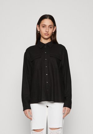 Black long-sleeve shirt with a collar, button front, and two chest pockets. The fabric appears smooth. Worn with ripped white jeans.