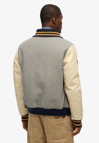 Chaqueta de cuerpo de lana gris con mangas de cuero crema, cuello de canalé azul marino con rayas amarillas y puños elásticos. Detalle destacado del logo en la manga.