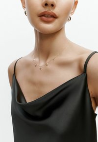 Arany nyaklánc öt geometriai medállal, amelyet fekete selyem camisole-lal viselnek, drapált dekoltázzsal és vékony pántokkal.