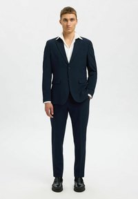 Marinblå kostym med en skräddarsydd kavaj, notch-lapels och två framfickor. Matchas med en vit skjorta med krage och svarta skor. Slät tygtextur.