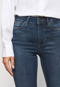 Jean en denim bleu foncé à coupe droite, avec une fermeture à bouton, cinq poches et des détails de surpiqûres contrastants.