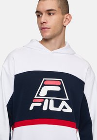 Witte en marineblauwe hoodie met een groot Fila-logo, rode accentstreep en een capuchon met trekkoord. Katoenmix met een gladde textuur.
