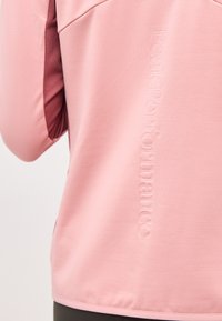 Κοντινό πλάνο στην πίσω πλευρά ενός απαλού ροζ σακακιού με ανάγλυφο κάθετο λογότυπο "peak performance" και ορατή λεπτομέρεια από πιο σκούρο ροζ ύφασμα στη μασχάλη.