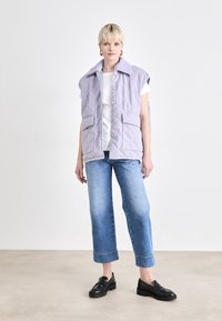 Lilafarbene, gesteppte Weste mit Kragen, Knopfverschlüssen und zwei vorderen Taschen. Kombiniert mit einem weißen T-Shirt und blauen Jeans mit weitem Bein.