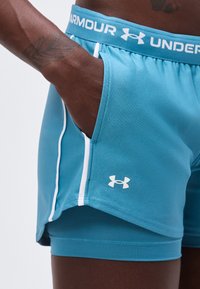 Άτομο με σκουρόχρωμο δέρμα που φοράει τιρκουάζ σορτς Under Armour με λευκή επένδυση, χέρι στην τσέπη και ορατό τατουάζ στον καρπό.