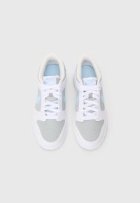 Pár bílých, šedých a světle modrých tenisek Nike se šňůrkami, z pohledu shora na jednobarevném světle šedém pozadí.