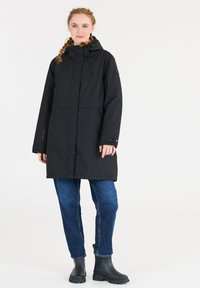 Svart parkas med dragsnodd på huvan, knappar framtill och rundad nederkant. Buren över blå jeans och svarta ankelkängor med texturerade sulor.