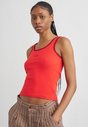 Kvinde med flettet hår iført en rød ærmeløs top og ternede bukser med den ene hånd i lommen mod en ensfarvet baggrund.