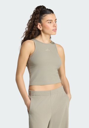 Joven con pelo rizado recogido en una coleta alta, que lleva un top corto sin mangas color topo y pantalones a juego con las manos en los bolsillos.