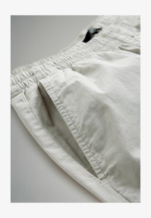 Svetlo bež hlače z elastičnim pasom, vidnim stranskim žepom, podrobnim šivanjem in mehakim teksturo tkanine na enobarvni podlagi.