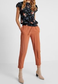 Blouse noire à motif floral avec manches courtes à volants, associée à un pantalon fuselé pêche doux et des bottines à talons beige.