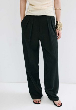 Femme portant un pantalon noir ample, un haut crème, des bracelets dorés et des tongs noires avec du vernis à ongles rouge, debout devant un fond blanc.