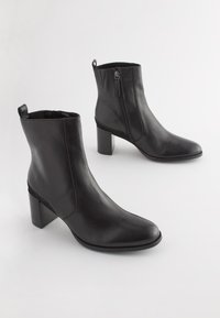 Bottines en cuir noir avec une texture lisse, un bout pointu et un talon carré. Dotées d'une fermeture éclair sur le côté et d'une boucle à l'arrière pour faciliter l'enfilage.