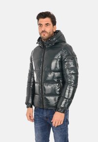 Giacca a vento verde scuro con una texture lucida e liscia, chiusura con zip, cappuccio e tasche laterali; presenta un motivo a righe orizzontali trapuntate.