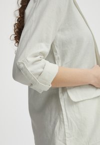 Helles Leinenhemd mit hochgekrempelten Ärmeln und Knopfdetails. Der Stoff hat eine glatte Textur und einen lässigen Schnitt.