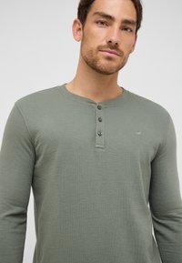 Zielona, długorękawowa koszula typu henley z teksturowanej tkaniny, z plisą na trzy guziki i małym logo na klatce piersiowej.