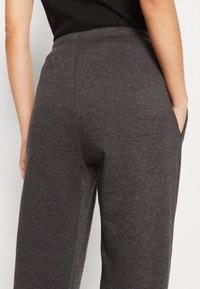 Person in dunkelgrauen Strickhosen und einem schwarzen ärmellosen Oberteil, mit den Händen in den Taschen, vom Taillenbereich bis zur Mitte des Oberschenkels gezeigt.