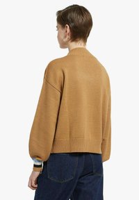 Donna con capelli corti castani che indossa orecchini a cerchio dorati, un maglione di lana color cammello con orlo e polsini a coste, e jeans blu scuro, vista da dietro.