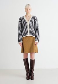 Striped black and white cardigan with a beige trim, worn over a brown suede skirt. Model wears knee-high brown leather boots.