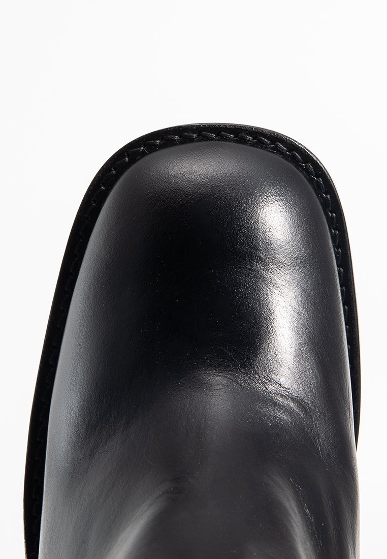 Bottine en cuir noir avec un bout arrondi, texture lisse et brillante, et surpiqûres contrastantes le long du bord. Design classique.