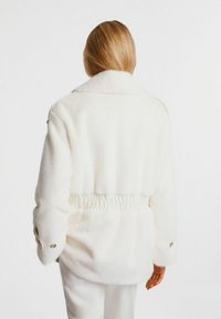 Biała polarowa kurtka z kołnierzykiem, elastycznym pasem i zapinanymi rękawami. Miękka tekstura, prosta forma i czyste linie.