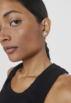 Boucles d'oreilles en perles accentuées d'or sur un modèle au teint lisse, portant un haut sans manches noir, montrant clairement les détails des boucles d'oreilles.
