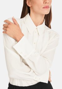 Weiße Bluse mit klassischem Kragen, langen Ärmeln und dezenten Falten. Auffällige Details sind quadratische Knöpfe an den Ärmelbündchen und eine weiche Textur.