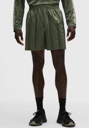 Hombre con pantalones cortos de rayas verdes, calcetines negros y zapatillas deportivas negras, de pie frente a un fondo claro y liso.