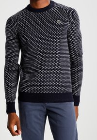 Pull en maille bleu marine avec un motif texturé blanc, col rond et poignets côtelés. Petit accent logo vert sur la poitrine.