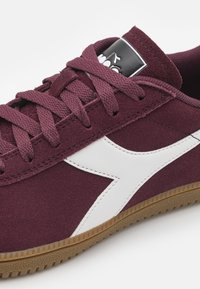 Burgundi velúr sneaker fehér díszítéssel és vastag fűzővel. A nyelvén fekete logópáttal és barna gumitalppal rendelkezik.